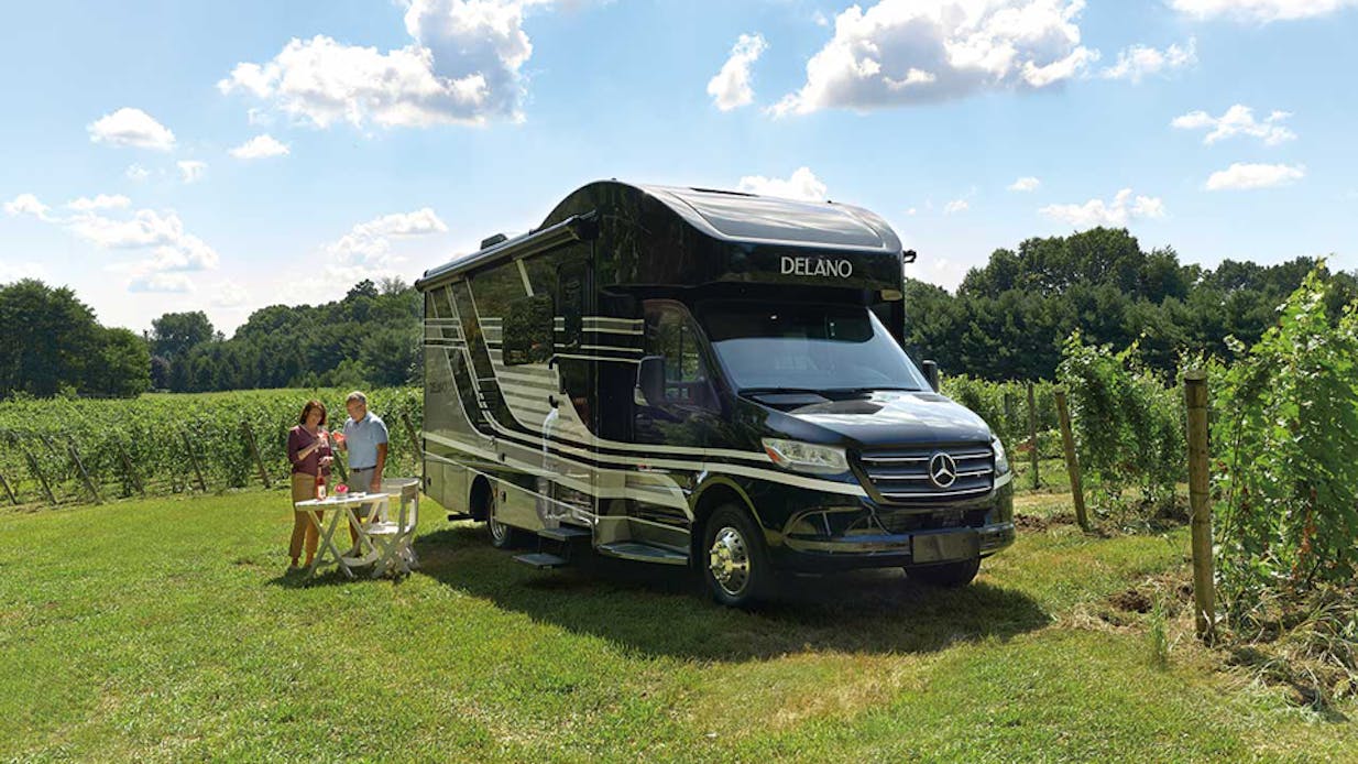 A couple standing next to their motor coach on a sunny day in a vineyard drinking wine