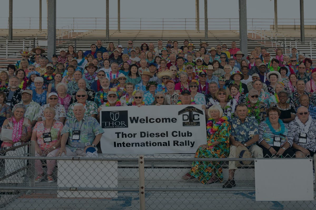 A group of motor coach owners sitting together with a large sign that says welcome Thor diesel club