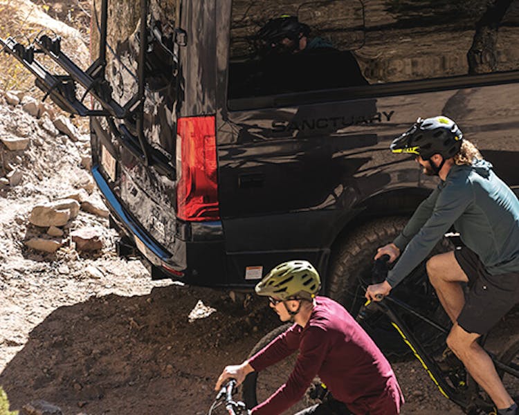 Two bicycle riders passing by their parked sanctuary sprinter van.